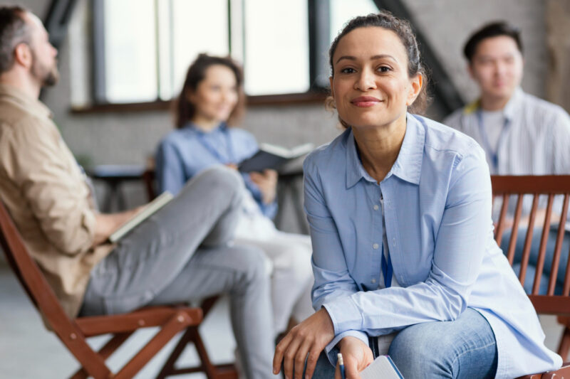 A woman in a light blue collared shirt sits in a room; three others are sitting behind her
