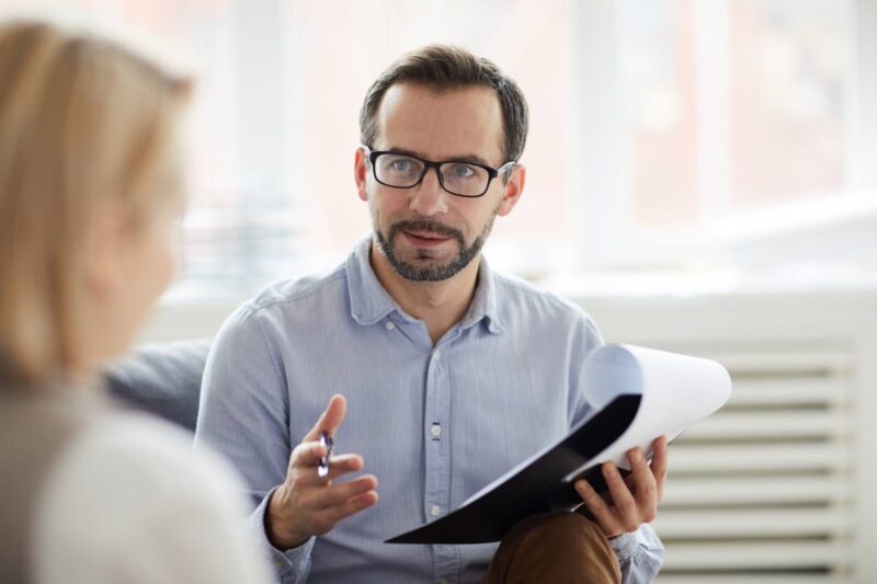 A man with a beard and wearing glasses holds paper and a pen and sits across from a woman